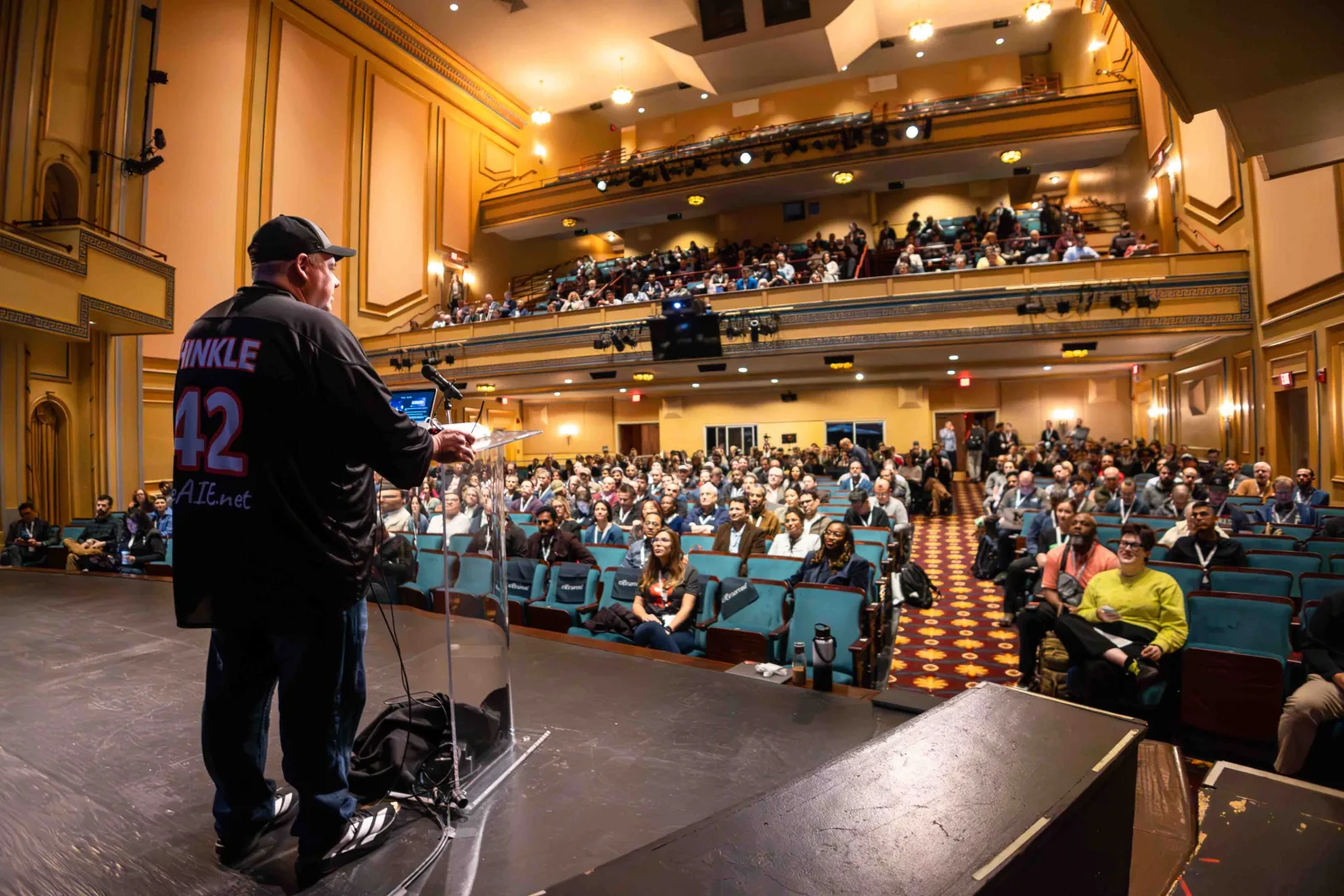 A speaker in a Hinkle 42 jersey addressing the full Carolina Theatre from the main stage at the 2025 conference